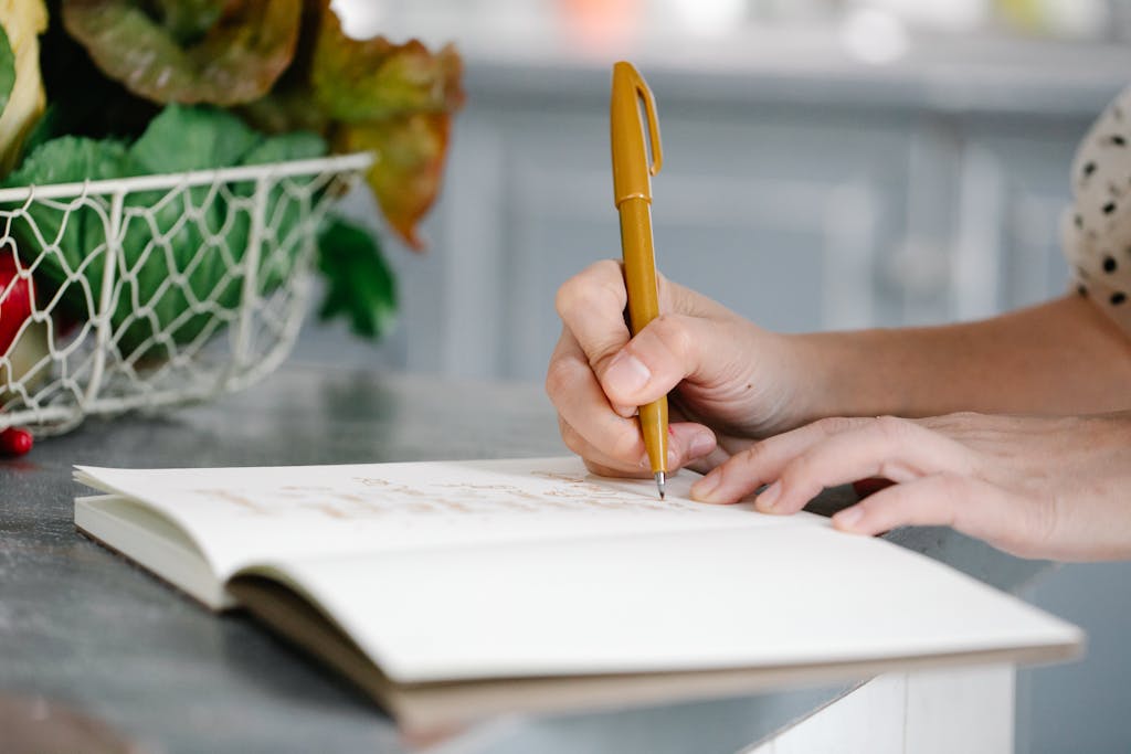 A woman writes in a notebook on a countertop near a basket of fresh produce.