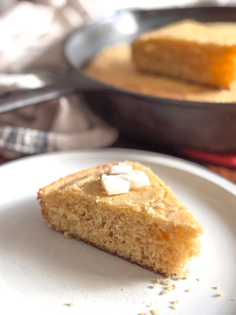 slice of cornbread on a plate with a cast iron skillet of cornbread in the background